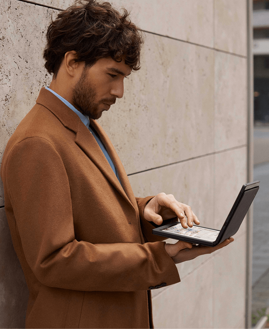 A man leaning against a wall holding a Lenovo ThinkPad X1 Fold open about 90 degrees in his hand