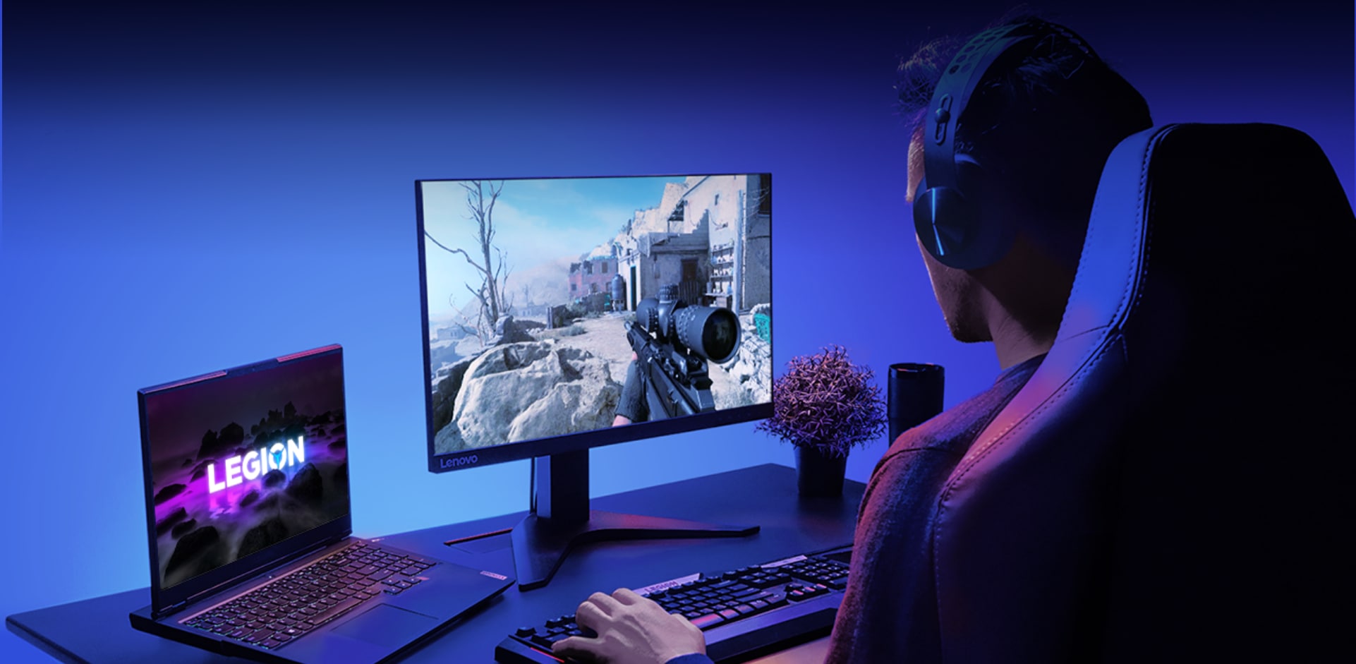 Геймер в наушниках играет в компьютерную игру, изображение которой видно на мониторе, сбоку расположен ноутбук