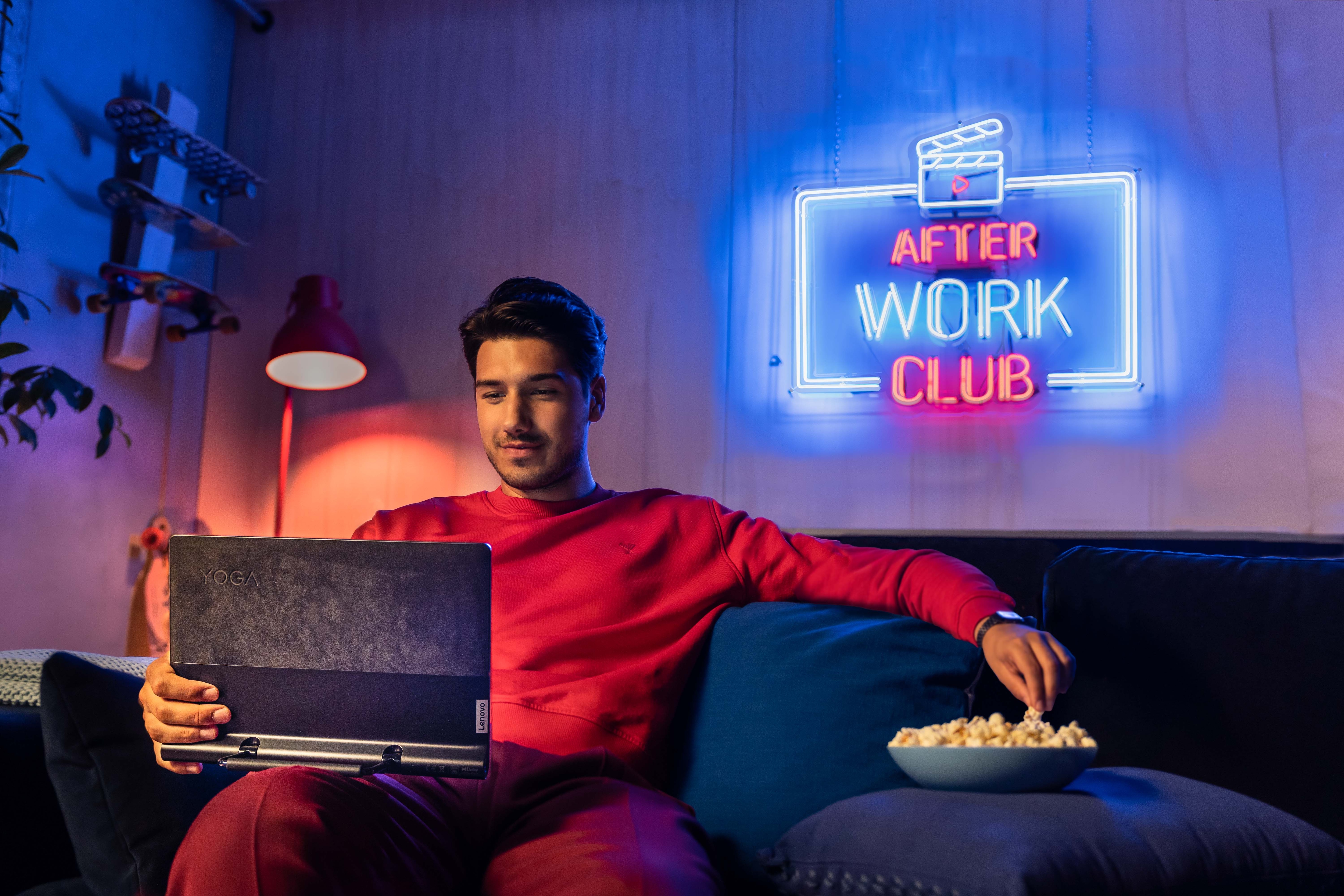 Homme assis sur le canapé avec un bol de pop-corn, en regardant une vidéo sur sa tablette Lenovo Yoga. Derrière le symbole du néon, il est lu « After Work Club ».