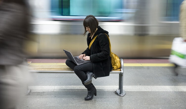 Young woman working on a Lenovo laptop on a subway bench with a moving train in the background