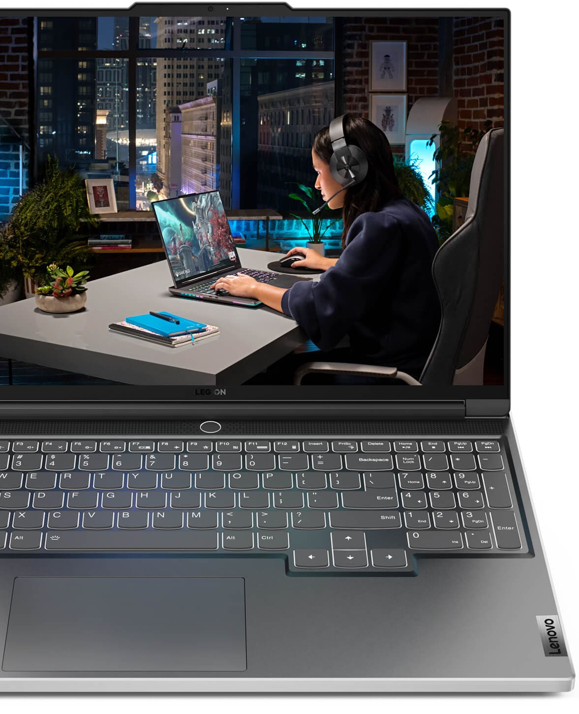 Lenovo Legion laptop with display showing image of young woman sitting at a table in an apartment room with windows overlooking a city at night as she plays a game on a Lenovo Legion laptop while wearing a Legion headset