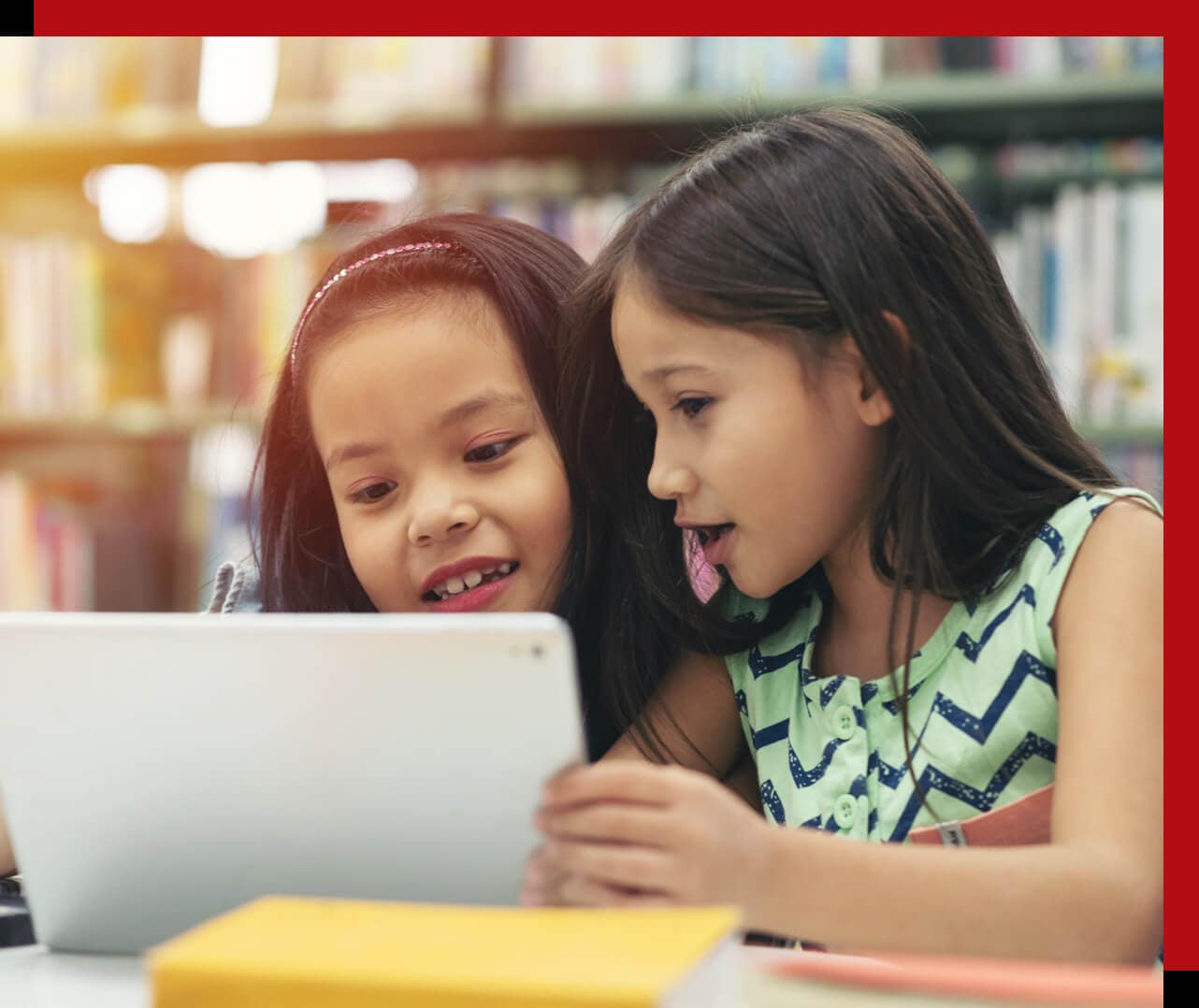 Two young girls at a table in a library, looking at the display of a Lenovo laptop or Chromebook