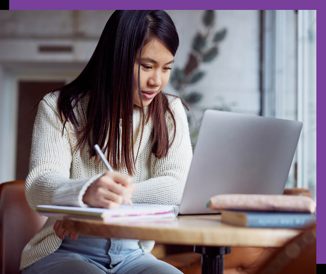 Young woman sitting at table at home, writing in a notebook while looking at something on the display of a Lenovo laptop