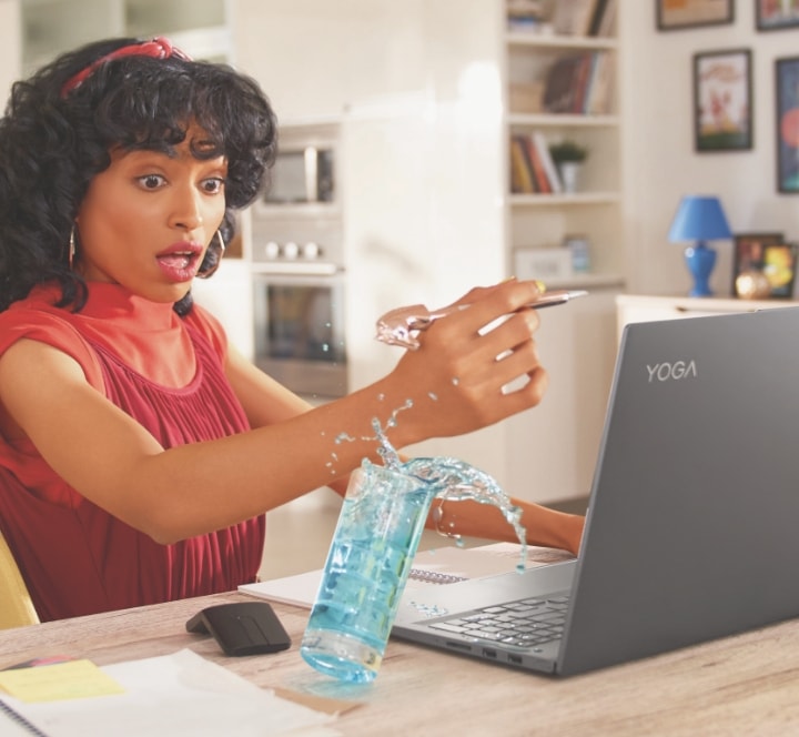 Woman sitting at a table working on her Lenovo Yoga laptop looking at a cup of water about to spill on her laptop